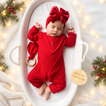 Load image into Gallery viewer, A sleeping newborn baby wearing a bright red ribbed one-piece outfit with a matching large bow headband and small matching hat, posed inside a white oval bassinet. The scene is styled with warm fairy lights, pine branches with red berries, and a round wooden sticker that reads 'My first Christmas.
