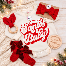Load image into Gallery viewer, A flat-lay holiday gift set for baby on a light wood background with a knitted blanket and fairy lights. The set includes a cream-colored long-sleeve bodysuit with a red and white 'Santa Baby' graphic, a red fabric bow headband, a wooden teething ring with a red bow, a kraft paper gift envelope with a red bow, and a round wooden plaque that reads 'my 1st Christmas'.