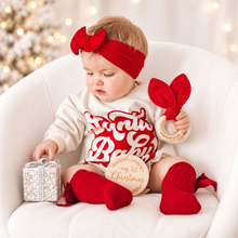 Load image into Gallery viewer, A baby sits in a white armchair wearing a red bow headband, a cream and red Christmas-themed long-sleeve shirt, and red knit socks. The baby holds a wooden teething ring with a red fabric bow and a round wooden plaque that reads 'my 1st Christmas'.
