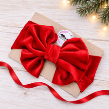 Load image into Gallery viewer, A large red velvet bow with matching red ribbon, tied to a kraft paper card with a round sticker, photographed on a light wood surface with pine branches and warm string lights in the background.
