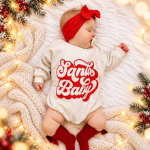 Load image into Gallery viewer, A baby wearing a cream long-sleeve bodysuit with a red 'Santa Baby' graphic, a red bow headband, and red socks, lying on a white blanket with holiday lights and pine branches.
